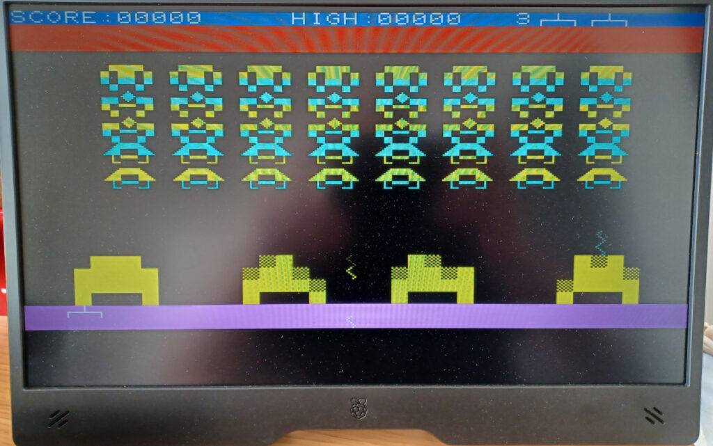 A game of space invaders being played on the Pico MZ (MZ-700). The aliens are a mixture of yellow and cyan. The score is 0 with a high score of 0. 3 bases are left.